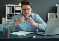 Man in blue shirt holding pens at desk with laptop.