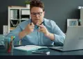 Man in blue shirt holding pens at desk with laptop.