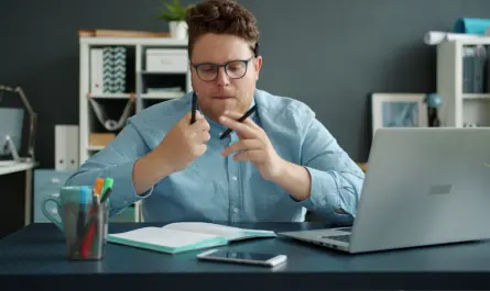 Man in blue shirt holding pens at desk with laptop.