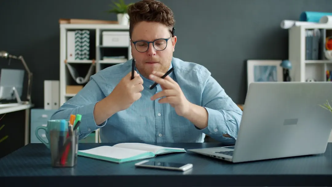 Man in blue shirt holding pens at desk with laptop.